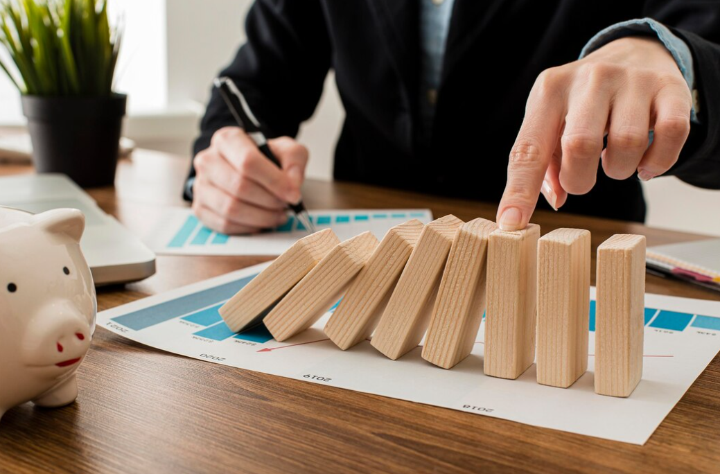 A person's hand on wooden dominoes on a table with charts, another hand makes notes