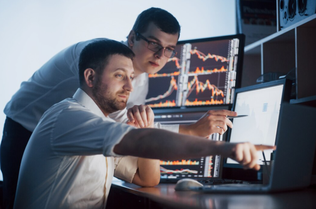 two men in white shirts, one is sitting at the table and pointing at the computer screen