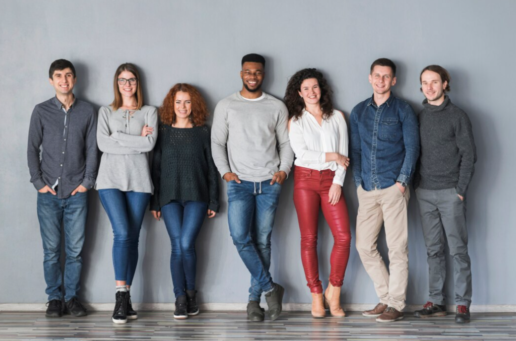 Seven people of varied genders and races standing side by side against a gray wall